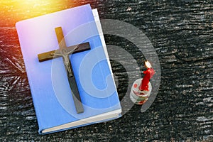 Closeup of wooden Christian cross on bible, burning candle and prayer beads on the old table.