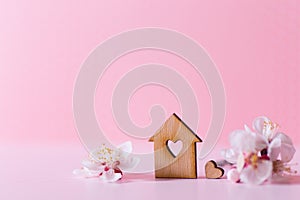Closeup wooden house with hole in form of heart surrounded by white flowering tree branches on pastel pink background