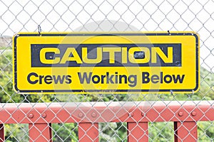 Closeup of a yellow [Caution Crews Working Below] sign on a chain-link fence under the sunlight