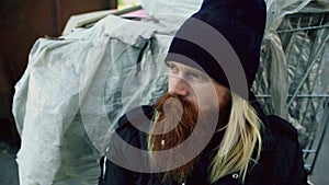 Closeup of young bearded homeless man sitting near shopping cart upet and stressed alone in the street
