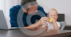 Closeup young bearded man sitting at desk at home with laptop Cute baby sits at high baby chair, father holds teether