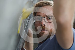 Closeup of young beautiful bearded caucasian man in glasses climbing on wall indoors during bouldering sport competition