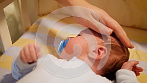 Closeup of young caring mother gently stroking her newborn baby boy lying in crib at night. Concept of happy parenting