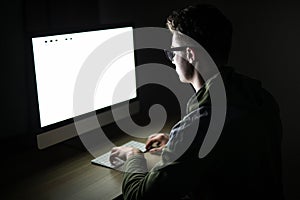 Closeup of young man sitting and using blank screen computer in dark room. hacker or programmer in dark room.