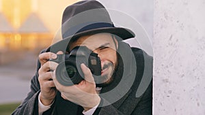 Closeup of young paparazzi man in hat photographing celebrities on camera while spy behind the wall