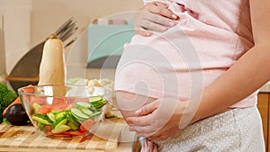 CLoseup of young pregnant woman cooking and eating vegetable salad holding big abdomen and touching it with hands