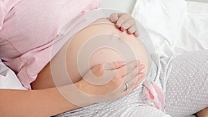 Closeup of young pregnant woman stroking her big belly while sitting on bed. Concept of parenting and happy expectation