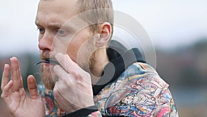 Closeup young smart man with beige beard playing on lips musical instrument in landscape of countryside, Steady cam