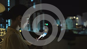 Closeup of young smiling woman texting on smartphone standing on rooftop terrace at night