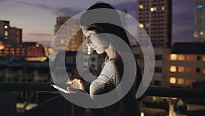 Closeup of young smiling woman texting on smartphone standing on rooftop terrace at night