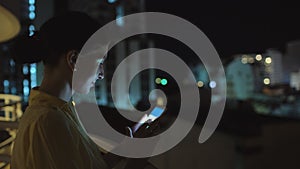 Closeup of young smiling woman texting on smartphone standing on rooftop terrace at night