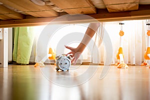 Closeup of young woman reaching under the bed and pressing snooze button on alarm clock