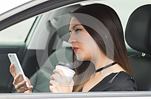 Closeup young woman sitting in car holding mobile phone and coffee cup, as seen from outside drivers window, female