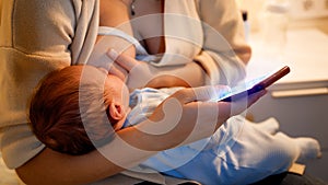 Closeup of young woman using smartphone and browsing internet while feeding her newborn baby son with breast milk at