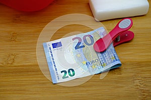 A clothes peg with a banknote, next to laundry soap and red basin, on a wooden plank. Housework bonus concept