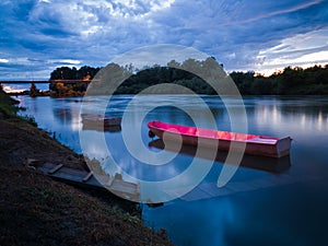 Cloud overcast evening during blue hour. Landscape with Sava river, moored fishing boats near wooden dock and bridge