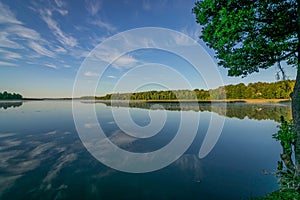 Cloud reflections in clear and calm lake water, forest in the background, summer morning