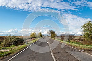 Cloudy country repaired highway/road in bad condition and blue cloudy sky. Green and yellow/orange fields, sunny landscape