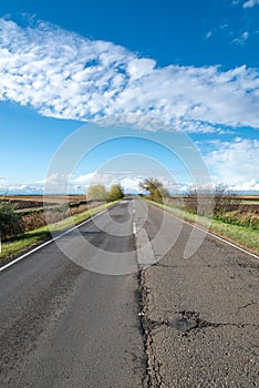 Cloudy country repaired highway/road in bad condition and blue cloudy sky. Green and yellow/orange fields, sunny landscape