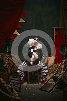 Clown in black hat, white face with red nose and striped pants performing over dark retro circus backstage background