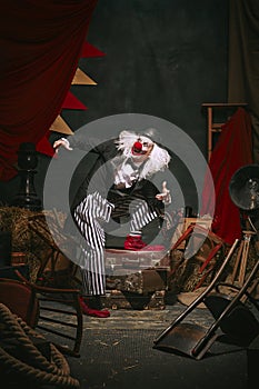 Clown in black hat, white face with red nose and striped pants playing over dark retro circus backstage background