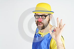 A clown in a bright blue and yellow suit, glasses and a hat shows an OK gesture