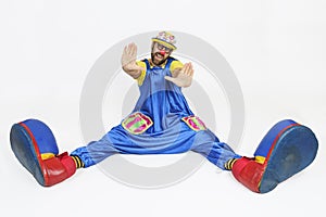 A clown in a bright costume sits with his legs apart and put his hands forward, palms