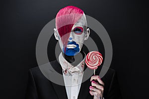 Clown and Halloween theme: Scary clown with pink hair in a black jacket with candy in hand on a dark background in the studio