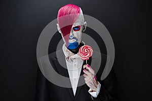 Clown and Halloween theme: Scary clown with pink hair in a black jacket with candy in hand on a dark background in the studio