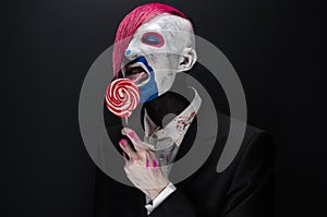 Clown and Halloween theme: Scary clown with pink hair in a black jacket with candy in hand on a dark background in the studio