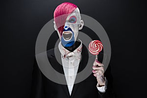 Clown and Halloween theme: Scary clown with pink hair in a black jacket with candy in hand on a dark background in the studio