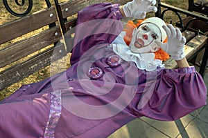 A clown with sharp teeth lies on a bench in a city park. The concept of fear and horror. Halloween or Carnival Cosplay for All