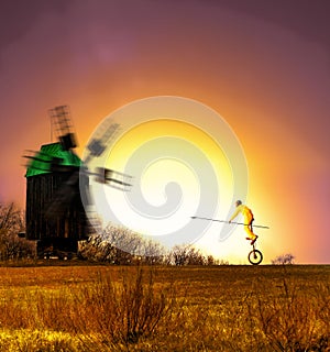 A clown on a unicycle riding with a lance in his hand towards the windmil during sunset photo