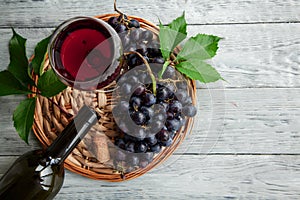 A cluster of dark grapes and wine glass, bottle on a round straw tray and grey wooden table. Wine composition flat lay