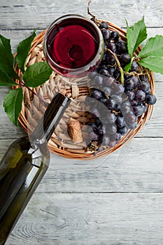 A cluster of dark grapes and wine glass with red wine, bottle on a round straw tray and grey wooden table. Wine composition flat