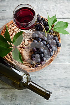 A cluster of dark grapes and wine glass with red wine, bottle on a round straw tray and grey wooden table. Wine composition flat