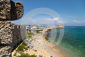 Coastline along the Mandraki  harbour in Rhodes city with gravel beach, crystal clear turquoise water, old windmills, Kastell