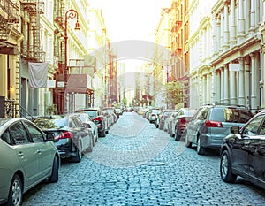 Cobblestone covered Greene Street is crowded with buildings and cars in the SoHo New York City with colorful light