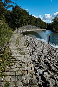 Cobblestone path near a waterway leading from a lake to a marina with a forest to the left. Vertical shot