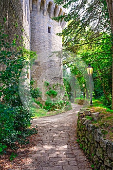 Cobblestone path with street light lamp near wall of stone brick medieval castle tower in green park in Republic San Marino