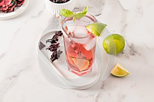 Cocktail glass with cold tea from hibiscus rose petals on a white tray and marble table. lime, mint leaves to improve the taste.