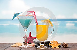 Cocktail and glass of orange juice with umbrellas, straws, starfish, smooth stones and sea shells on background of sea.