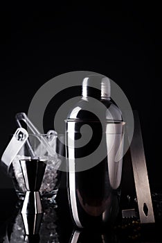 Cocktail shaker, swizzle, tongs and spoon with ice in a bucket for preparing a summer cocktail beverage on a black table