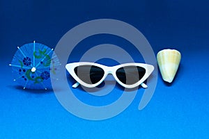 Cocktail umbrella, stilish white sunglasses and a shell on a bright blue background. The concept of summer sea vacation