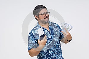 A cocky male tourist in a Hawaiian shirt flaunting money and a boarding pass, isolated on a white backdrop