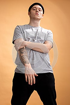 Cocky stylish guy dressed in a gray shirt, black jeans and black hat with tattoo on his hand is standing on the beige