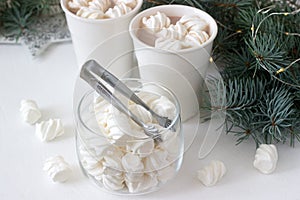 Cocoa with marshmelow in white cups and marshmallows in a glass jar on a white table with spruce branches and garland.