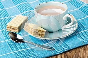 Cocoa with milk in cup, piece of wafer on saucer, wafer, spoon on napkin on table