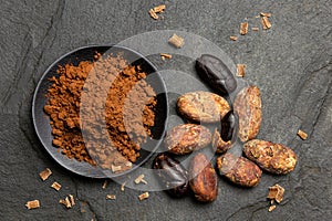 Cocoa powder in a black ceramic dish next to roasted peeled and unpeeled cocoa beans and chocolate shavings on black slate from