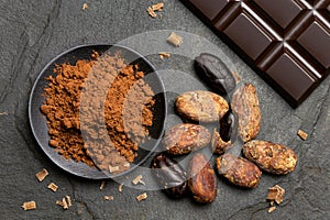 Cocoa powder in a black ceramic dish next to roasted peeled and unpeeled cocoa beans, chocolate shavings and a slab of dark